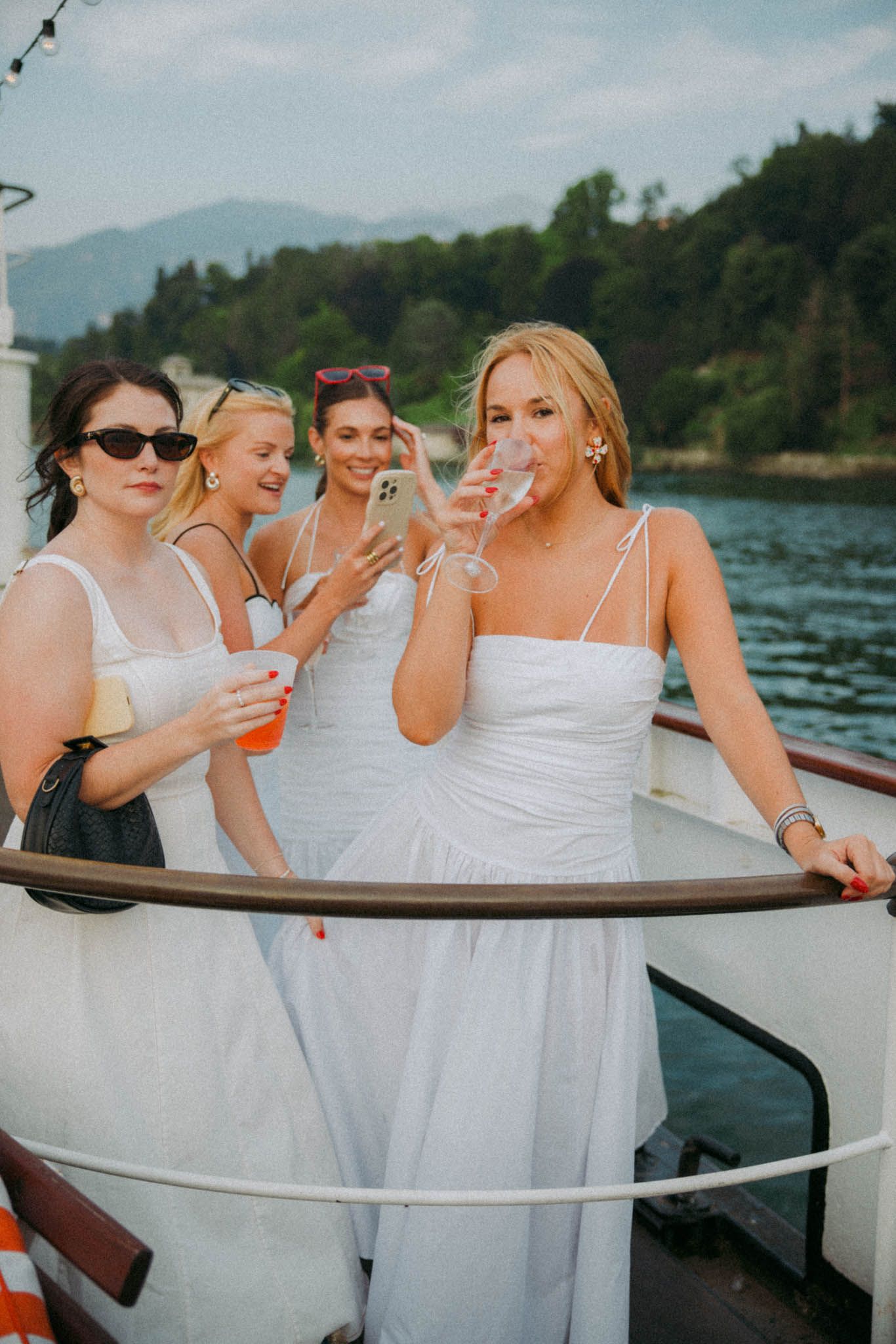 A group in white dresses laugh and socialize together on a boat with mountains visible in the background.