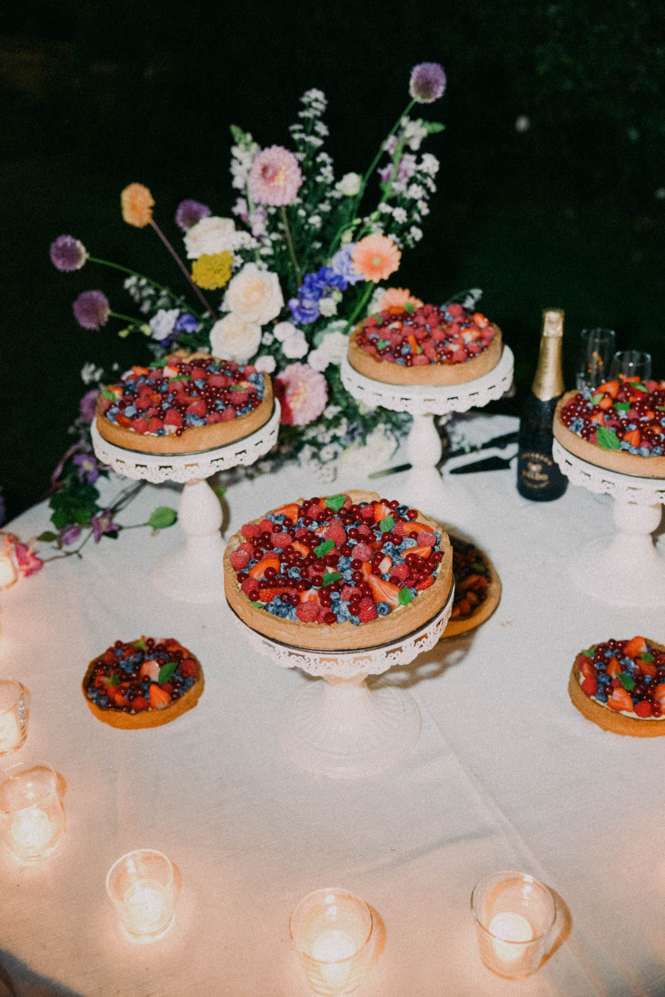 Wedding dessert table decorated with fresh berries and flowers features multiple tarts on cake stands with glowing candles.