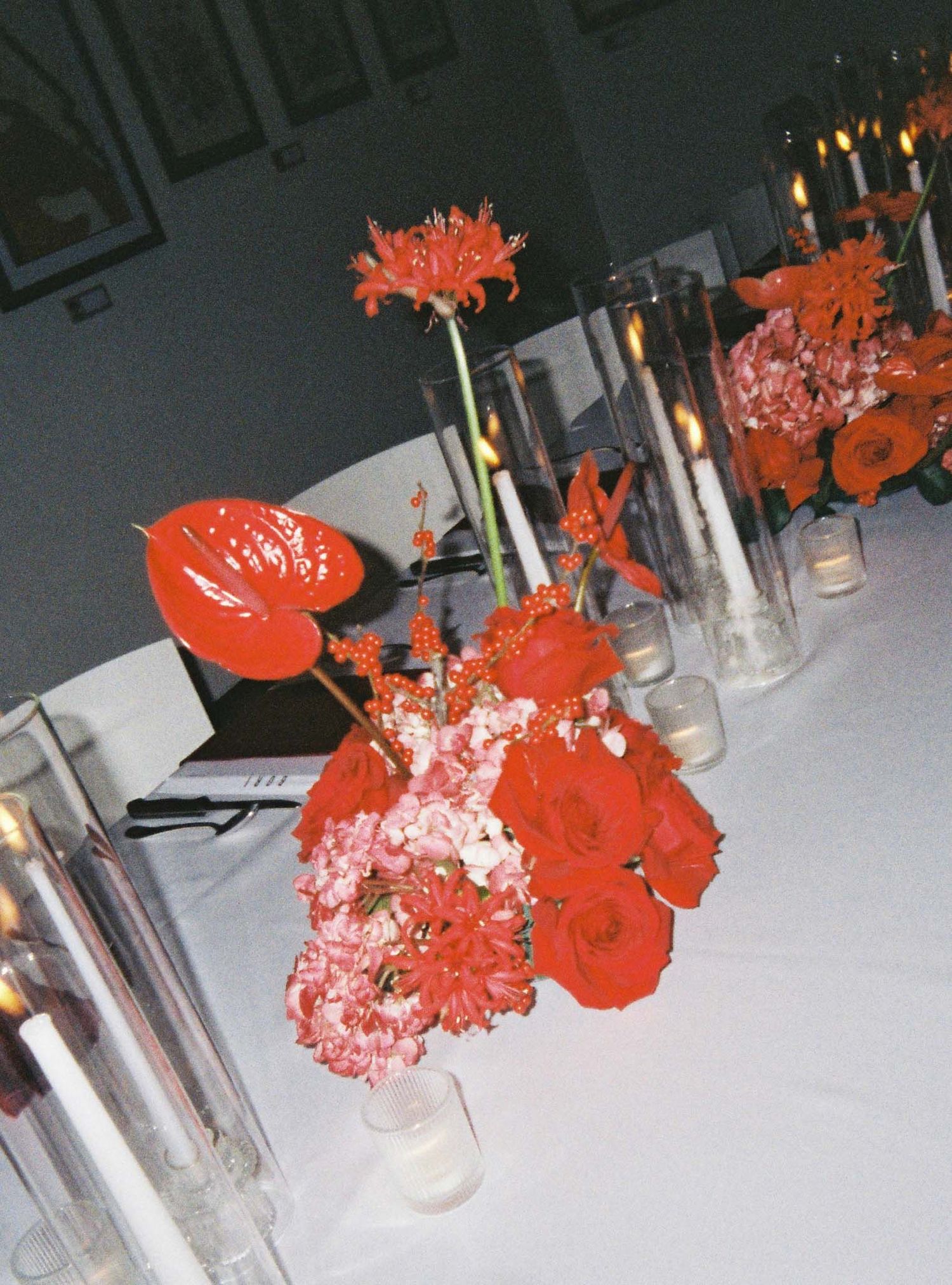A series of red floral arrangements on a table with candles create an elegant dining atmosphere.