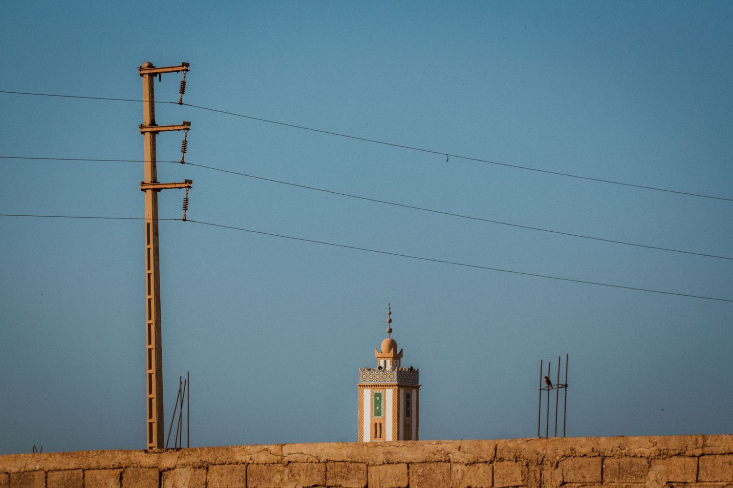 Minaret of a mosque rises against blue sky with electrical power lines and poles in Morocco.