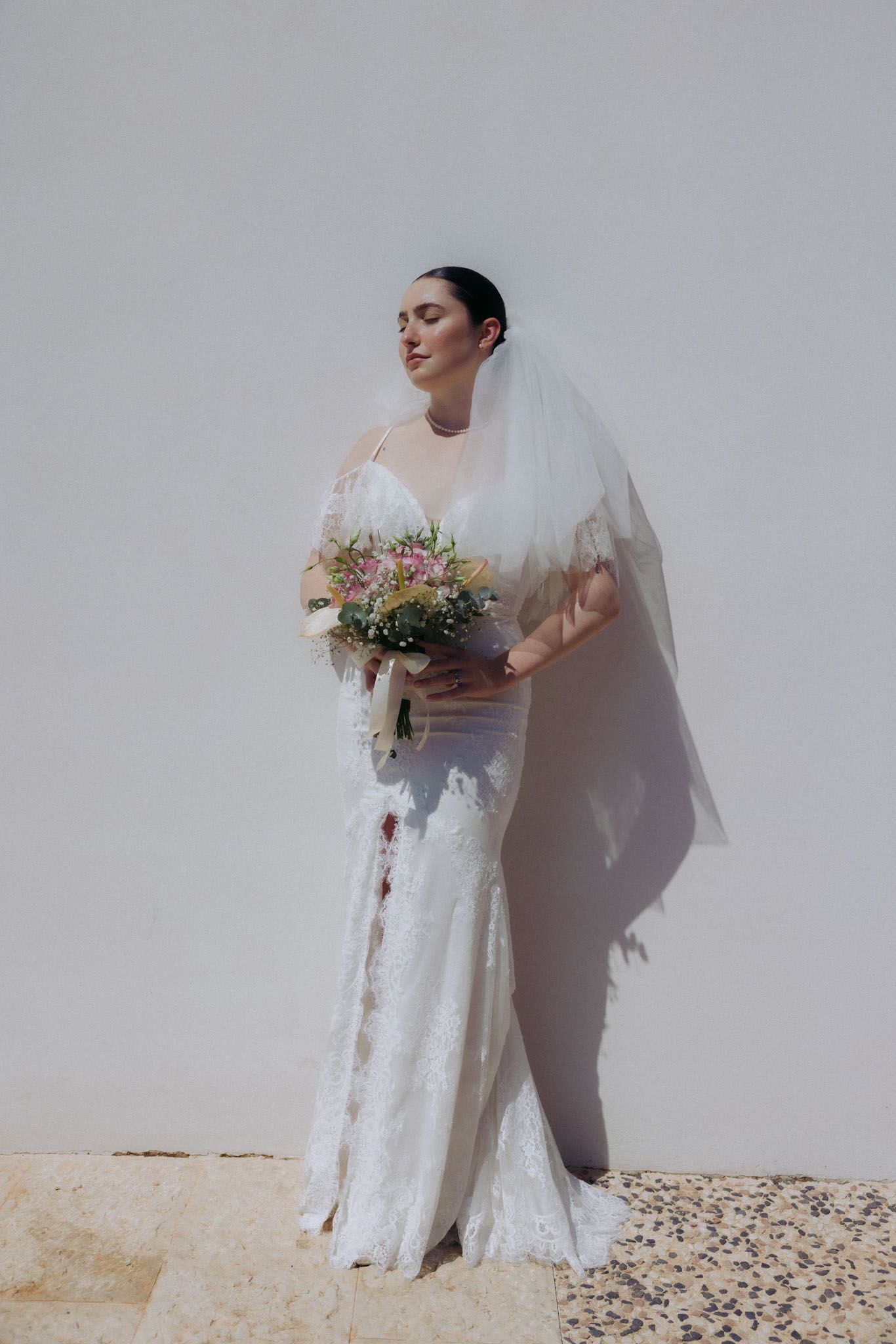 Bride in flowing white lace wedding dress and veil poses against light wall while holding colorful floral bouquet.