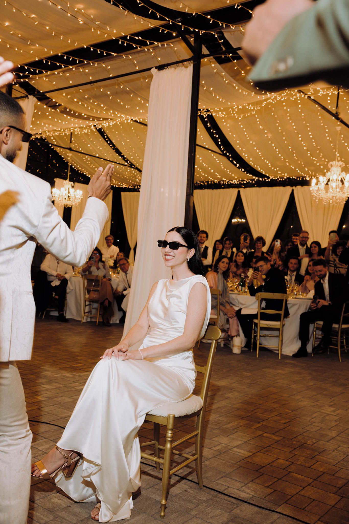 Wedding guests watch as someone performs a dance routine in a white suit on a wooden dance floor under twinkling lights.