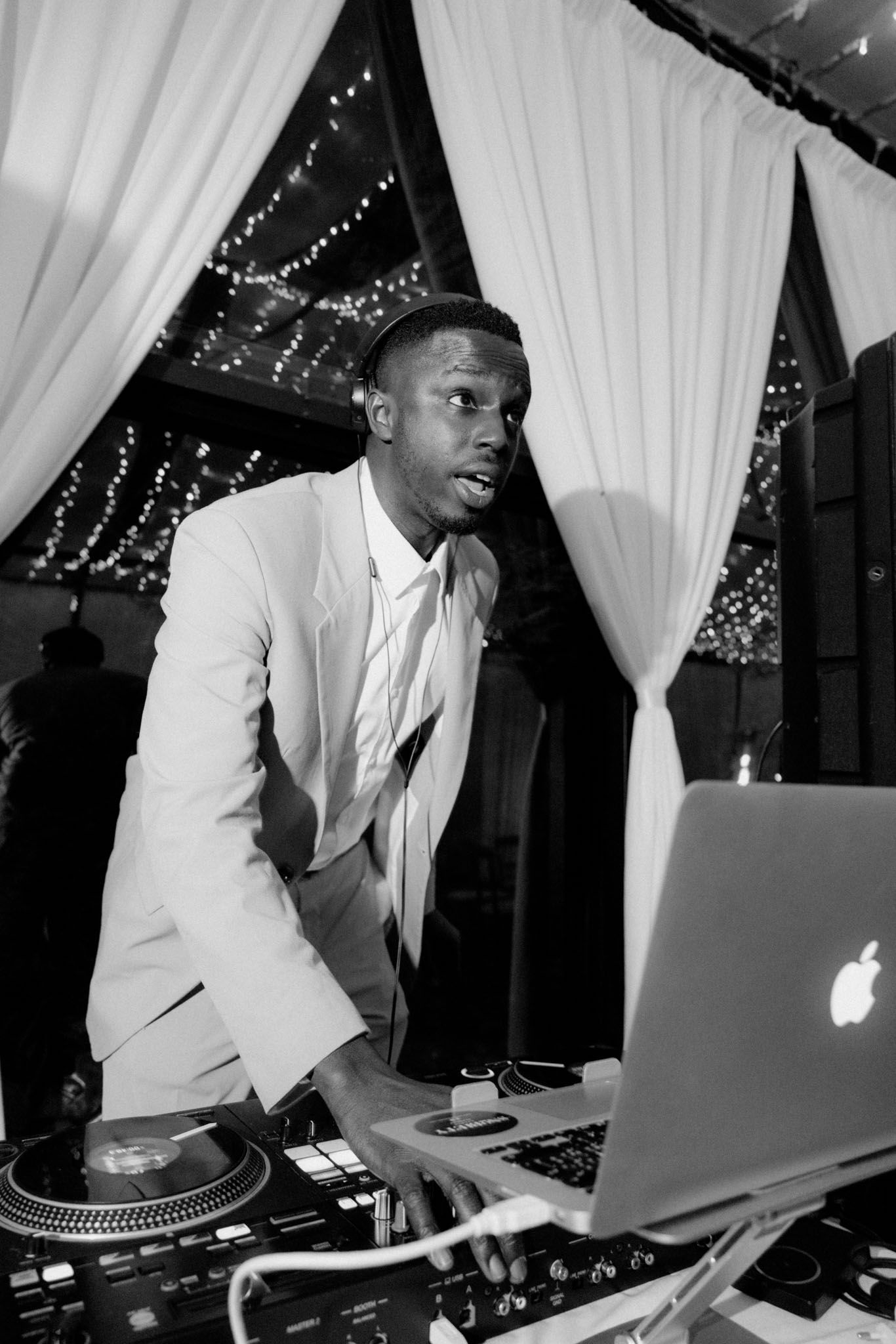 DJ performs at a nightclub using turntables and a laptop under white fabric draping and twinkle lights.