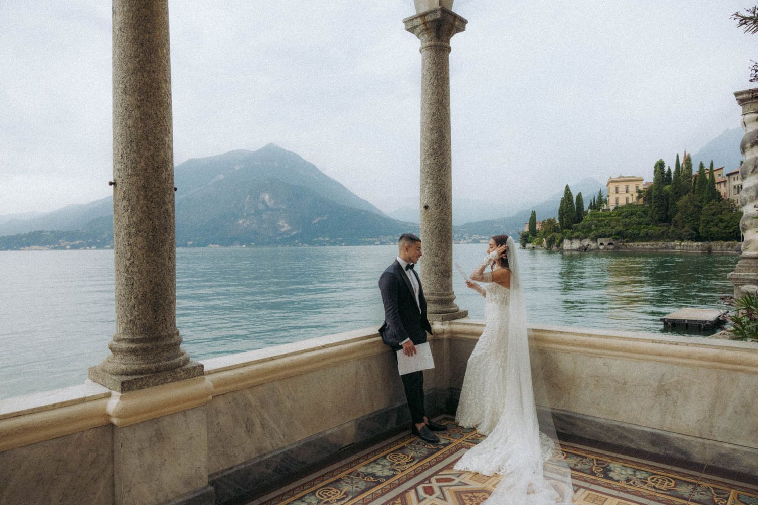 Wedding couple enjoys romantic views from an ornate stone balcony overlooking Lake Como in Italy.