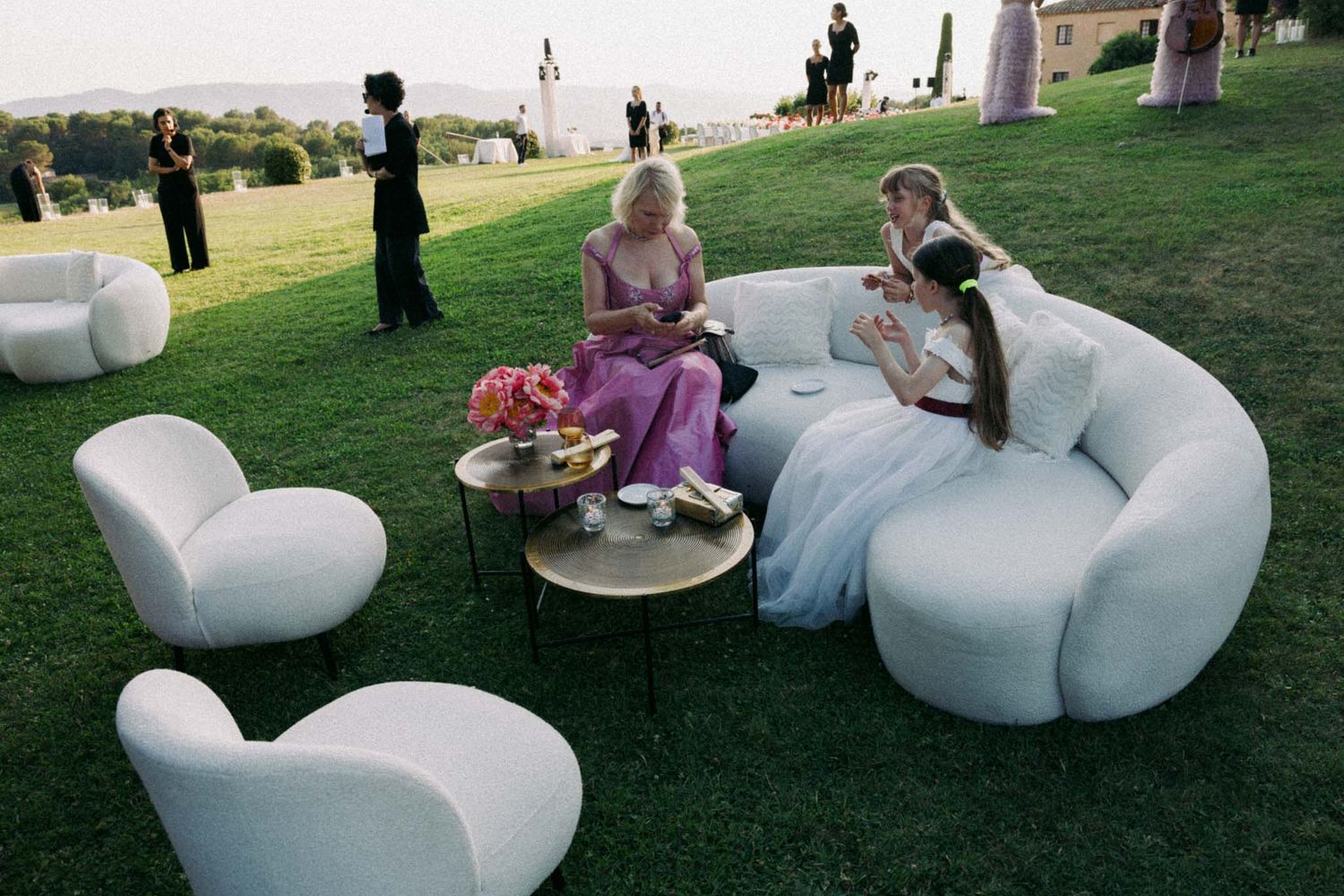 People relax and socialize on large white sculptural seating installations in a grassy outdoor park setting.