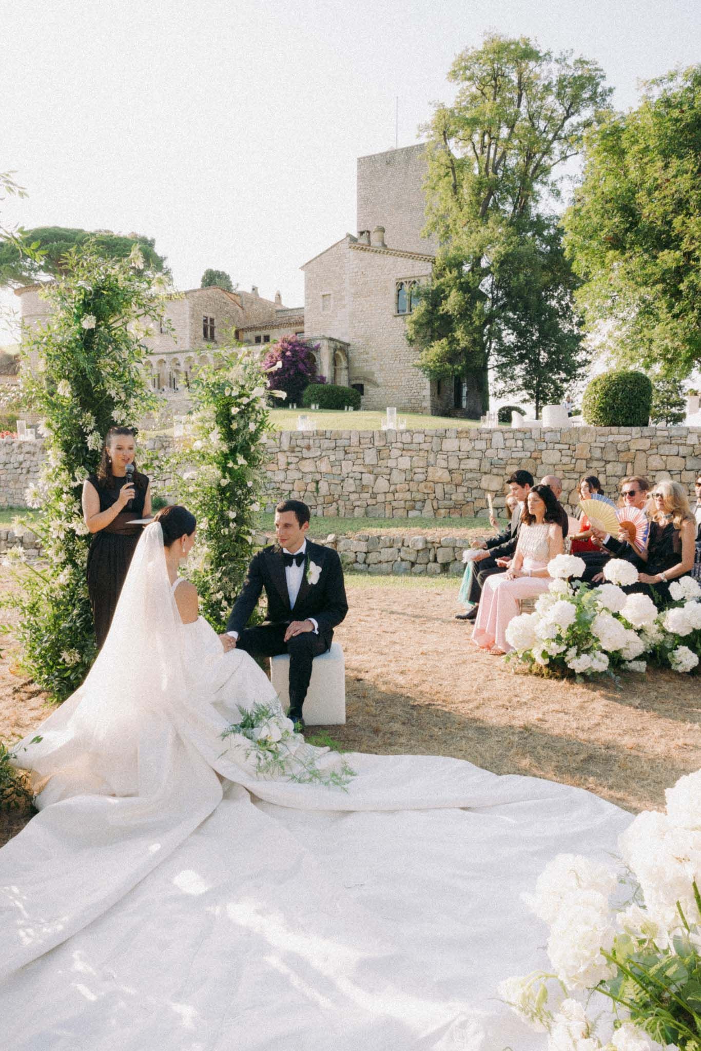 Elegant wedding couple poses in front of stone castle wall with floral arrangements and flowing white dress.