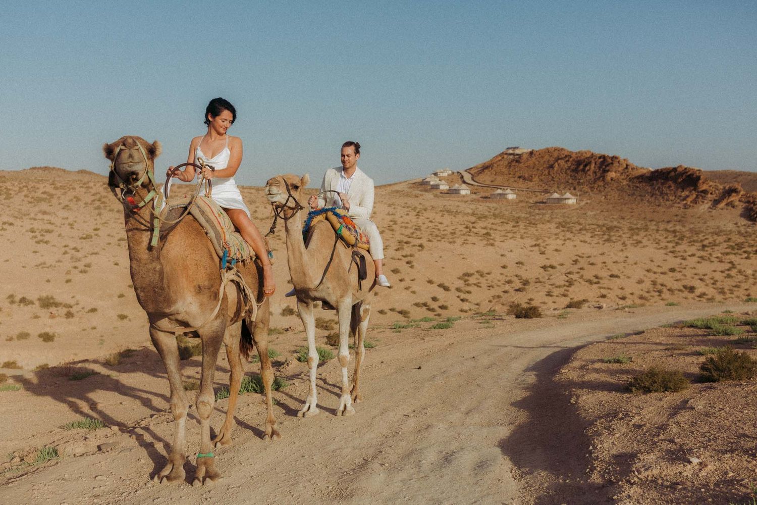 Two tourists ride camels through a desert landscape with sandy hills and clear blue sky in the background.