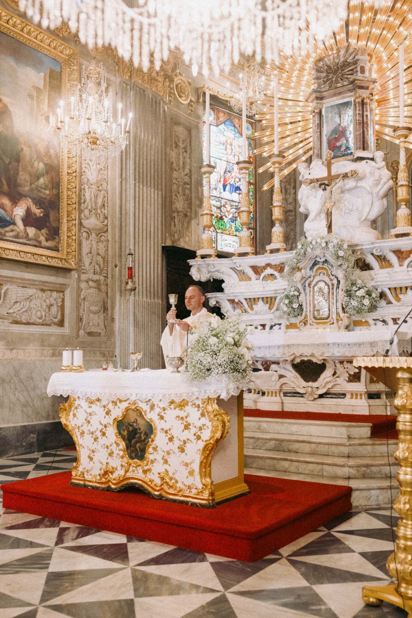 Ornate baroque church interior with crystal chandeliers, marble floors, and white floral decorations on red velvet platforms.