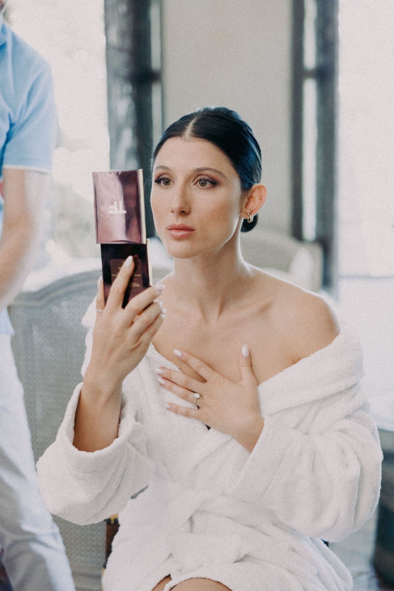 Someone in a white bathrobe holds a pink cosmetic product while getting ready in a bright interior setting.