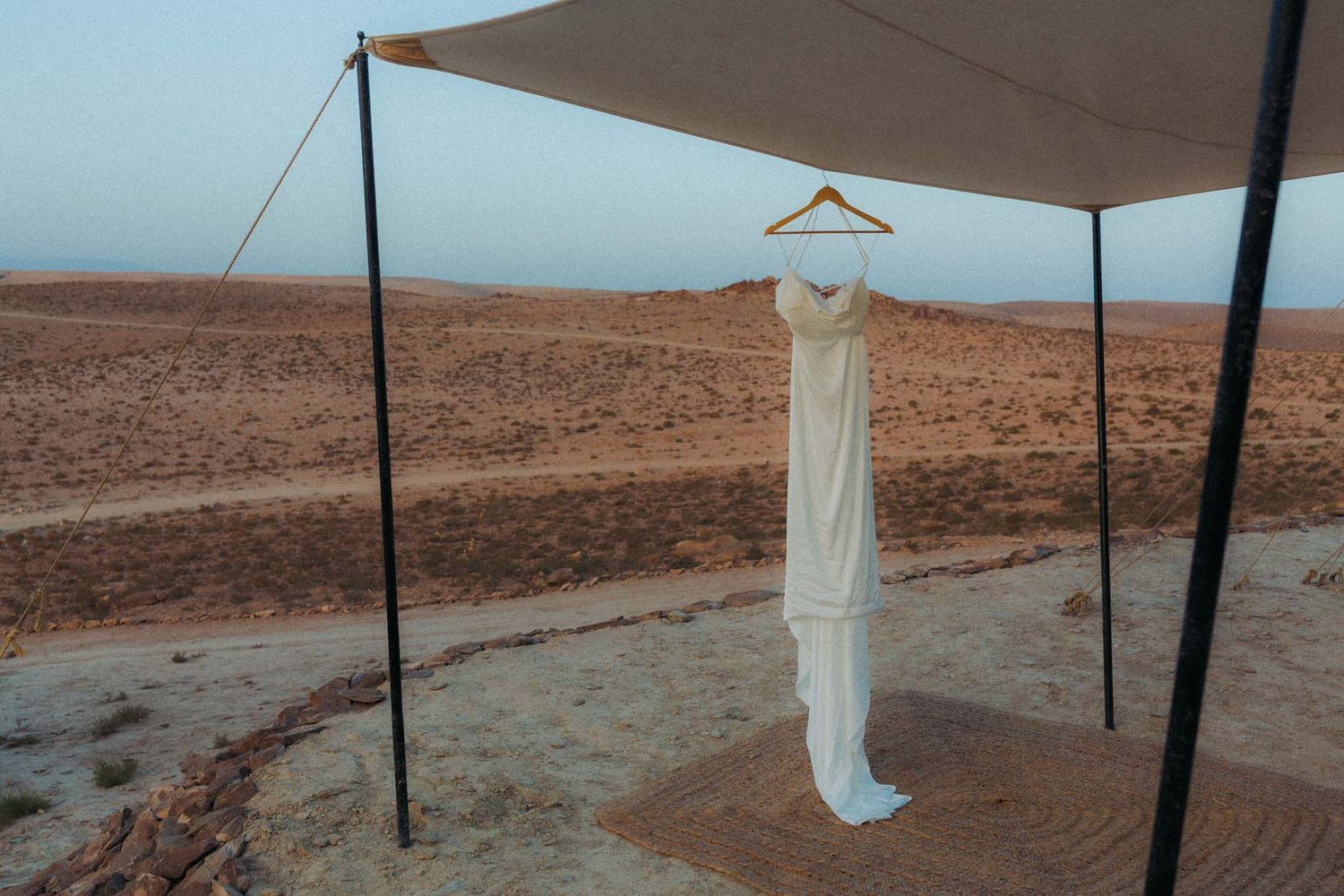 A white wedding dress hangs on display under a tan canopy tent in a desert landscape setting.
