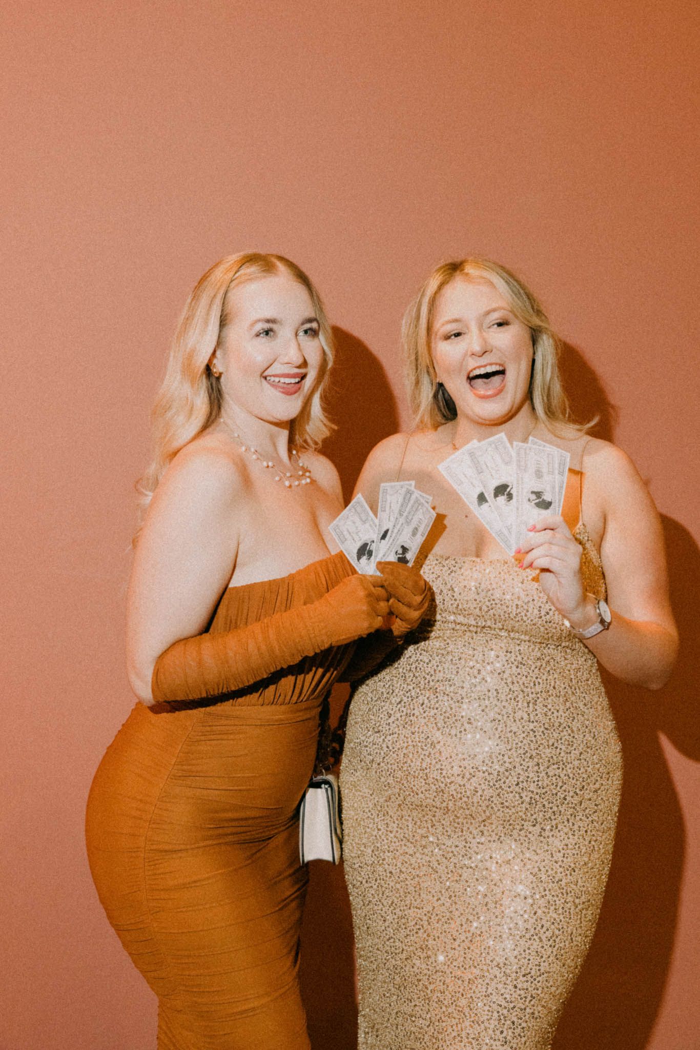 Two women in formal dresses laugh while holding playing cards at an event against a coral-colored background.