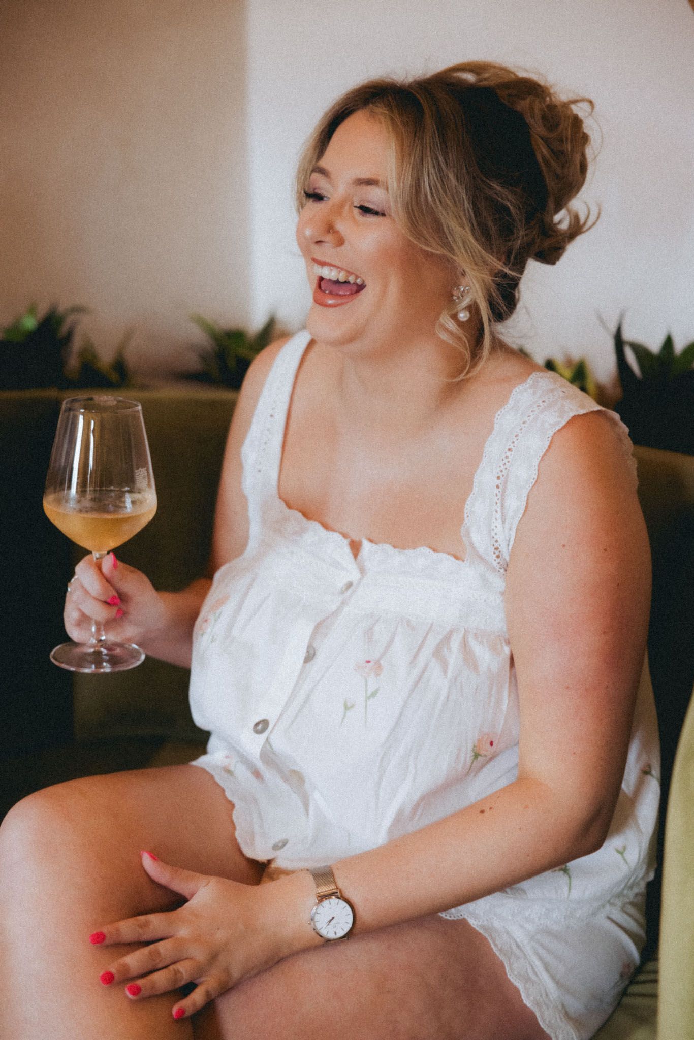 Person in white sleeveless dress laughs while holding wine glass in casual indoor setting.