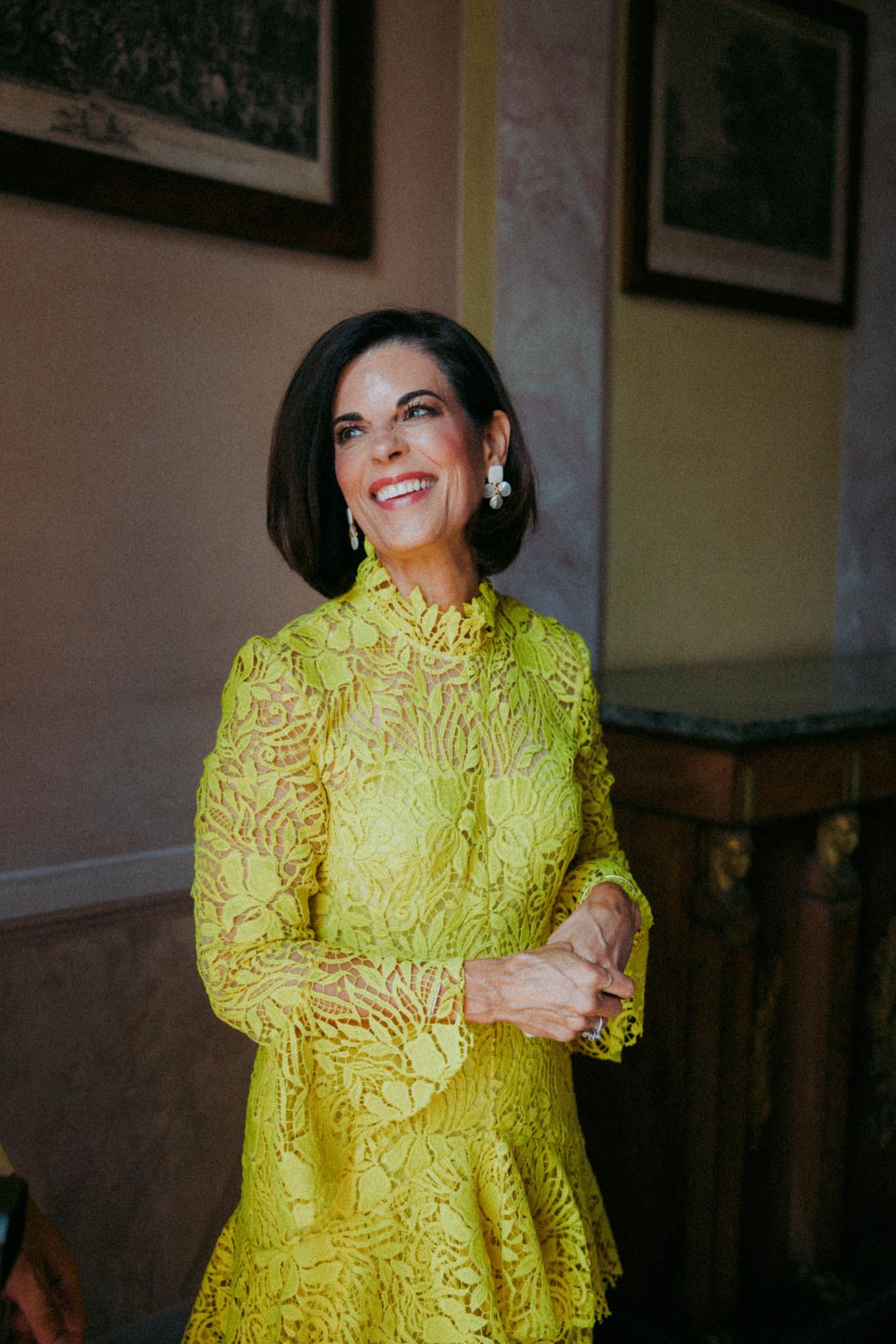 Someone wearing a vibrant yellow lace dress poses against a dark interior wall with framed artwork.