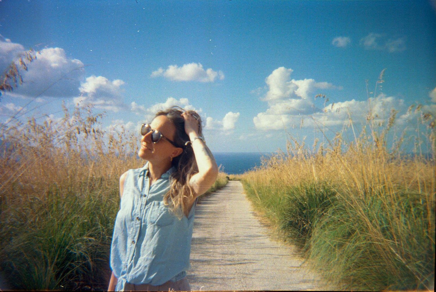 A person in a blue shirt walks along a sandy beach path through tall grass on a sunny summer day.