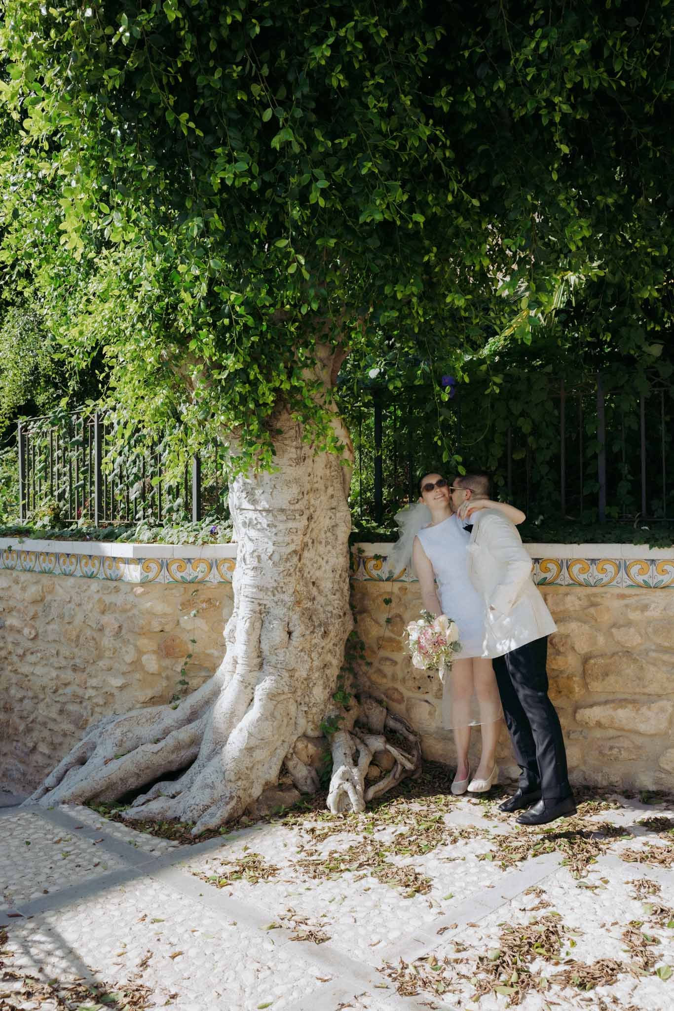 Romantic couple poses against stone wall and tree trunk in outdoor garden setting.