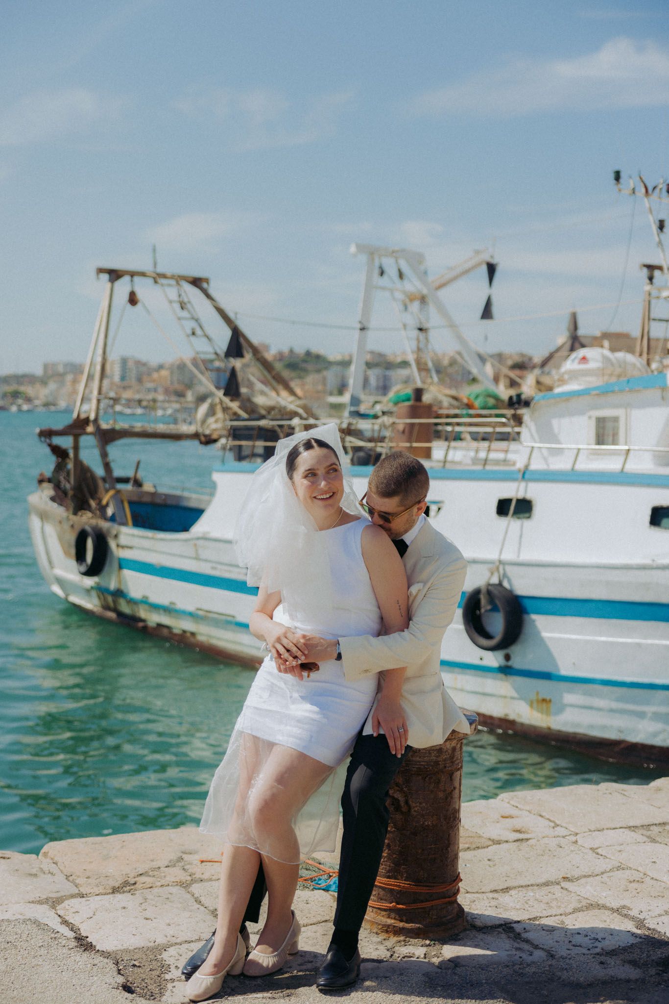 Couple sharing romantic moments by fishing boats docked at a Mediterranean harbor on a sunny day.