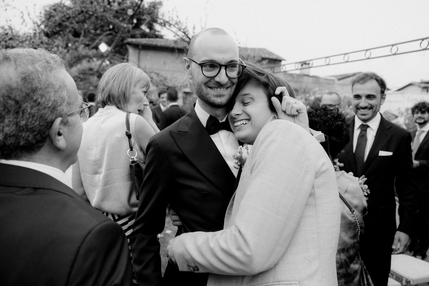 Black and white candid photo of wedding guests embracing and celebrating during an outdoor reception.