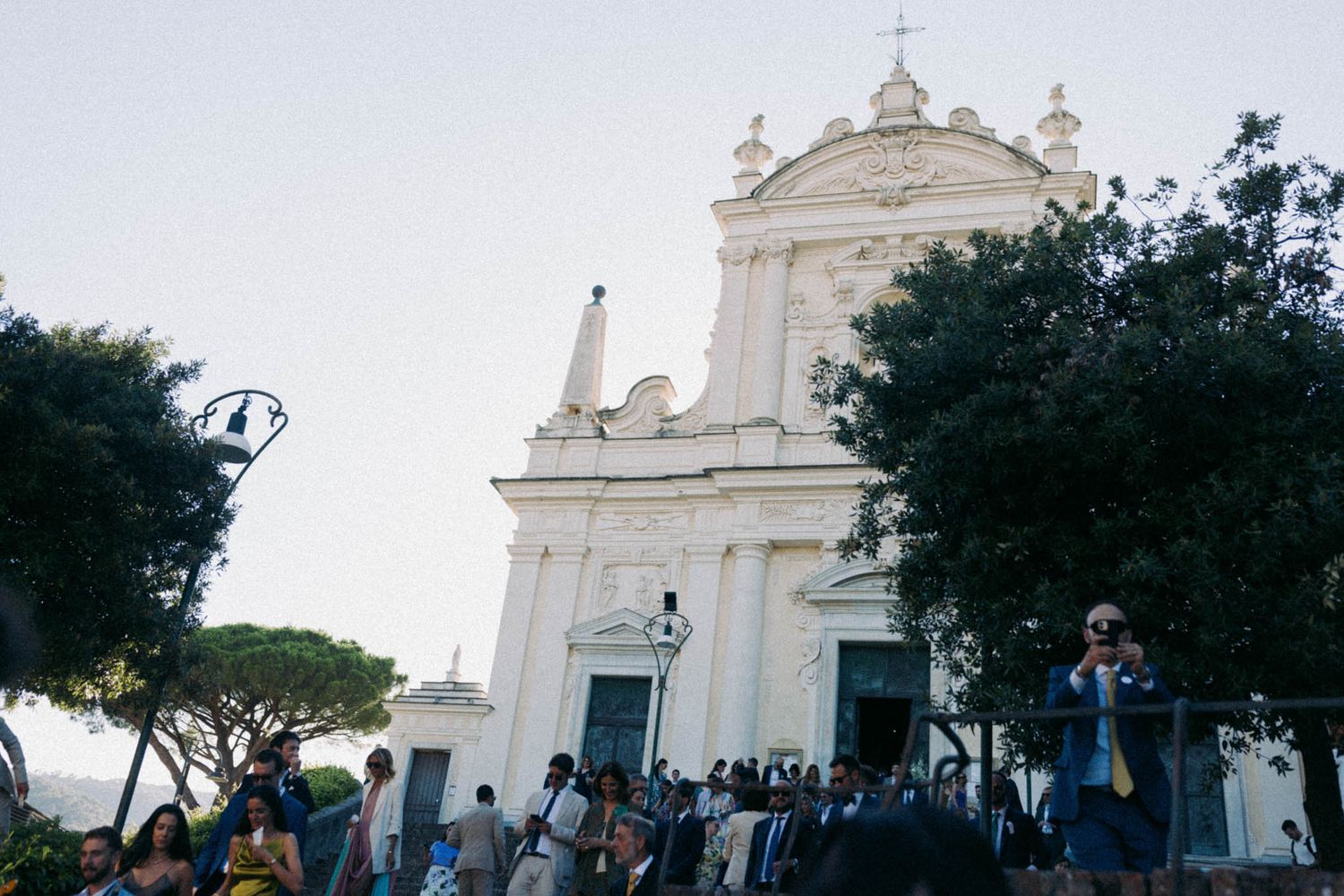 White church facade with cross and ornate architectural details stands against blue sky and trees in Italian setting.