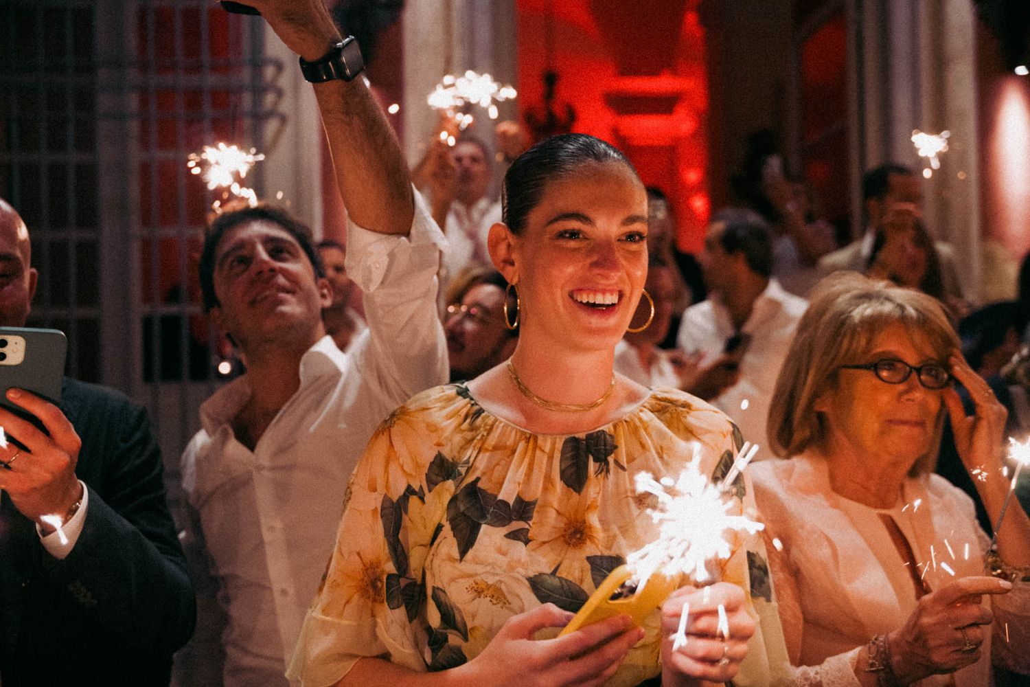 Wedding guests hold sparklers and smile during an evening celebration at an indoor reception venue.