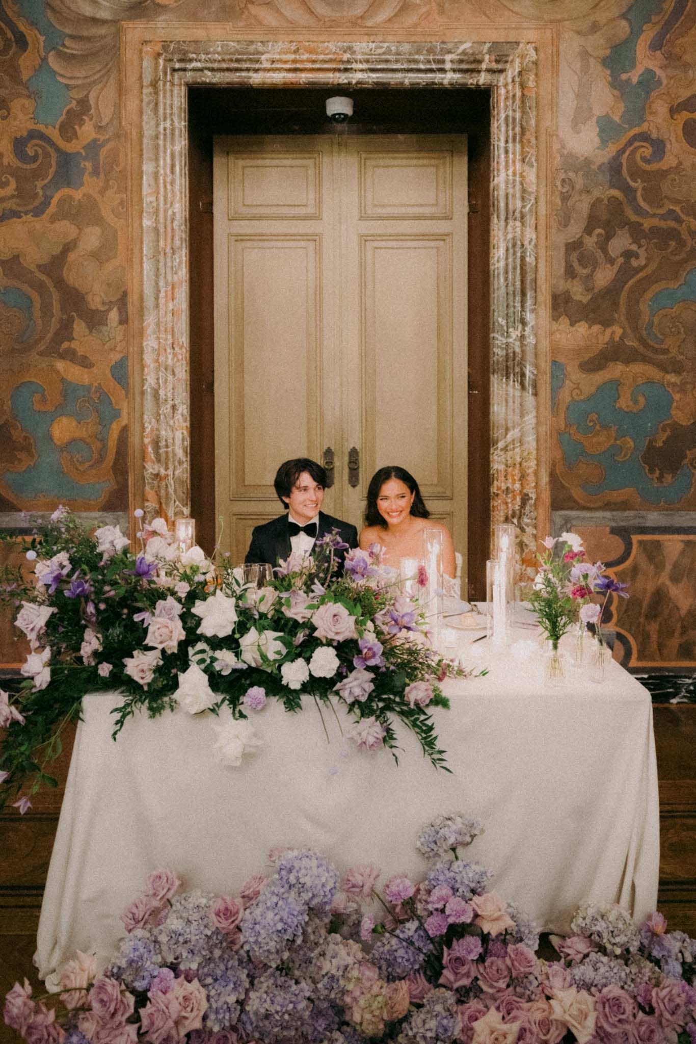 Elegant wedding head table with cascading purple and pink floral arrangements against ornate wallpapered backdrop.
