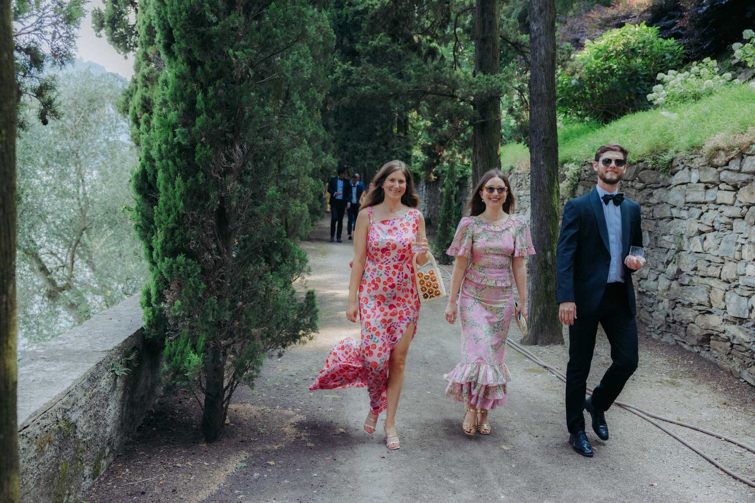 Wedding party walks along a scenic garden path surrounded by lush greenery and stone walls.