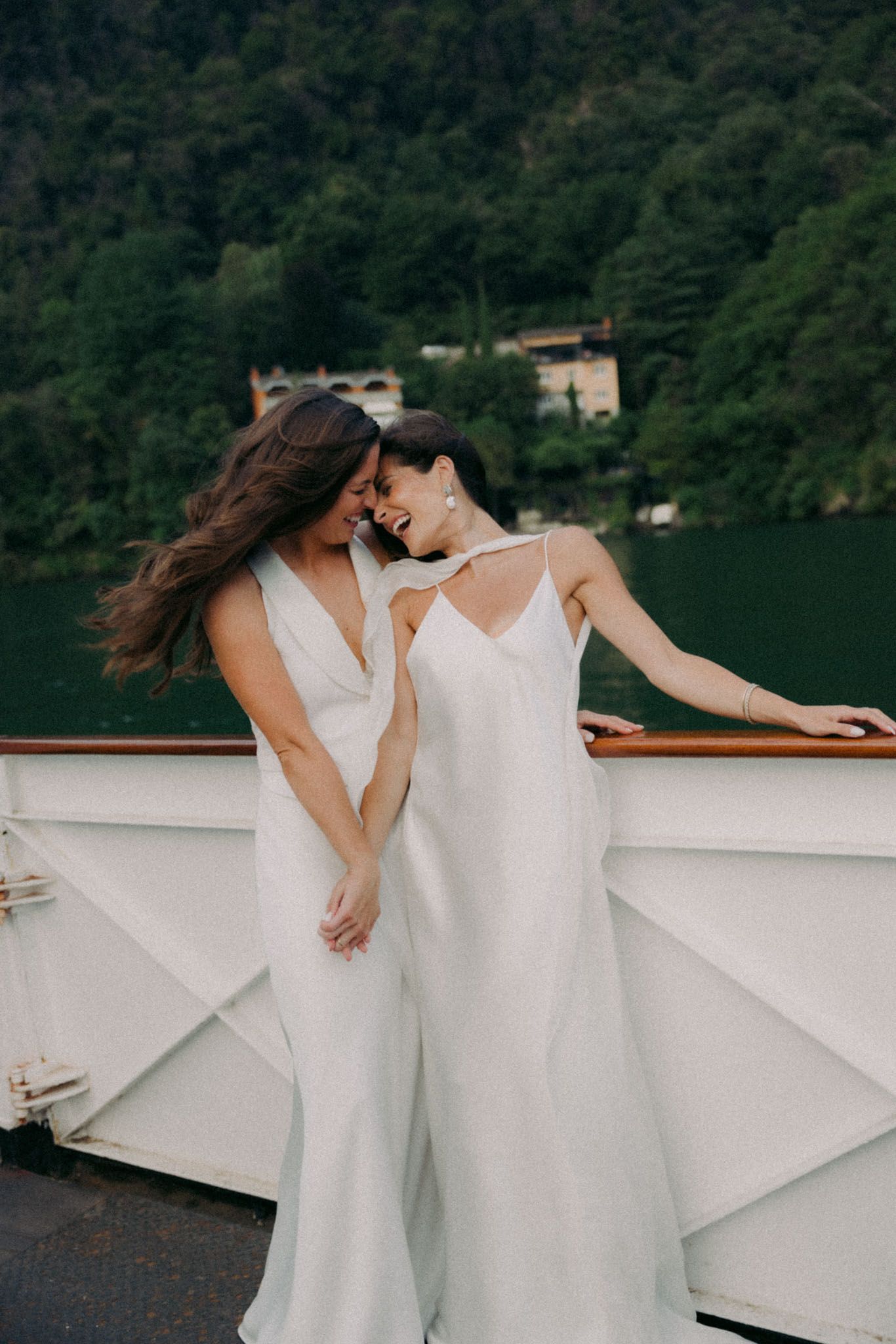 Two people in flowing white dresses dance together on a boat deck with a forested shoreline in the background.