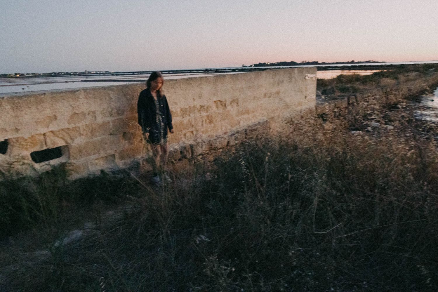 A person stands near a stone wall at dusk with dry grass and brush in the foreground.
