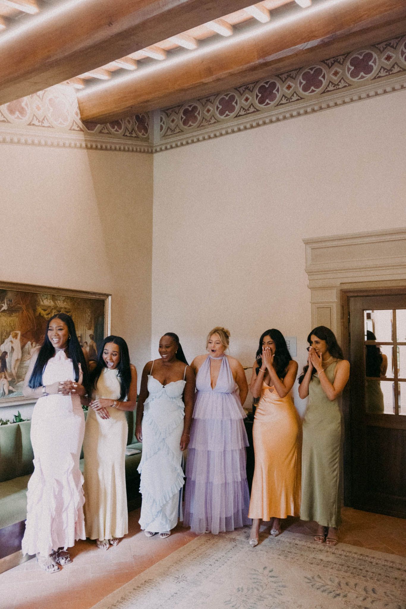 A group of bridesmaids in pastel-colored dresses stand facing a wall in a rustic indoor venue.