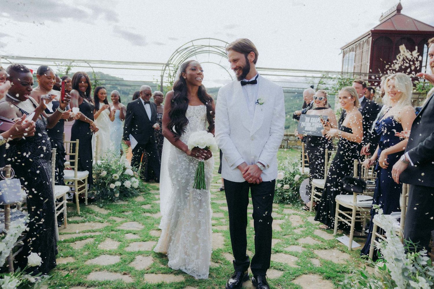 Wedding guests celebrate and throw confetti as newlyweds walk down the aisle at an outdoor ceremony.