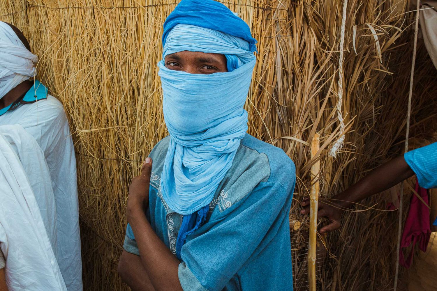 People wearing traditional blue and white desert clothing stand against a backdrop of golden straw or reeds.