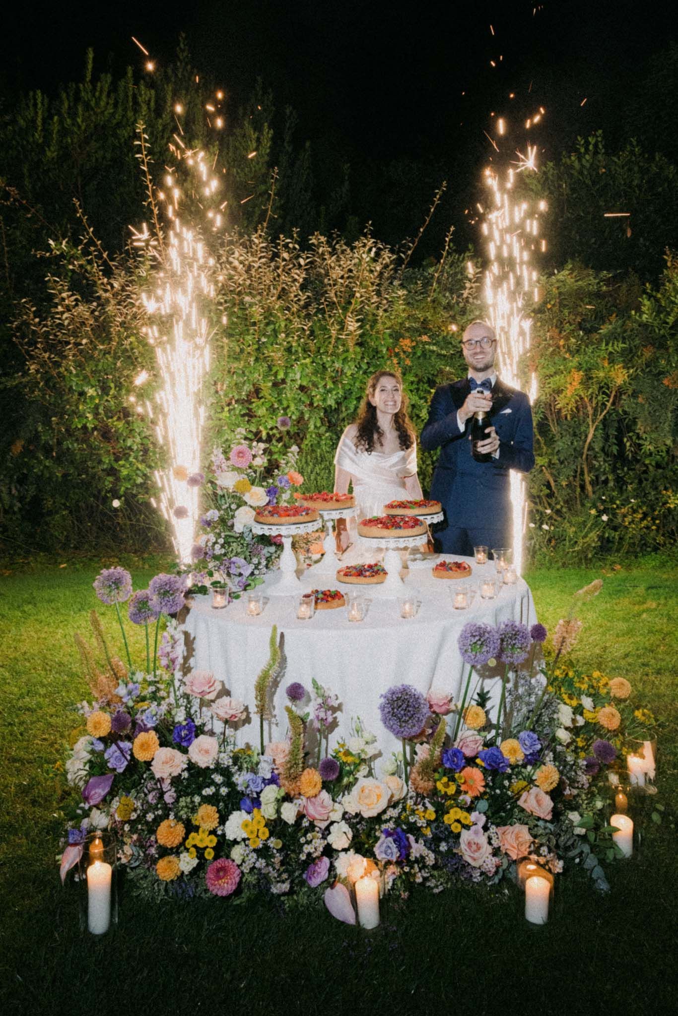Elegant wedding dessert table with sparklers and colorful floral arrangements in a garden setting at night.