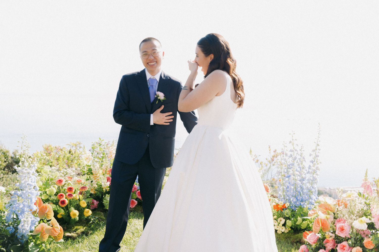 Couple shares an intimate moment during outdoor wedding ceremony surrounded by colorful flowers and ocean views.