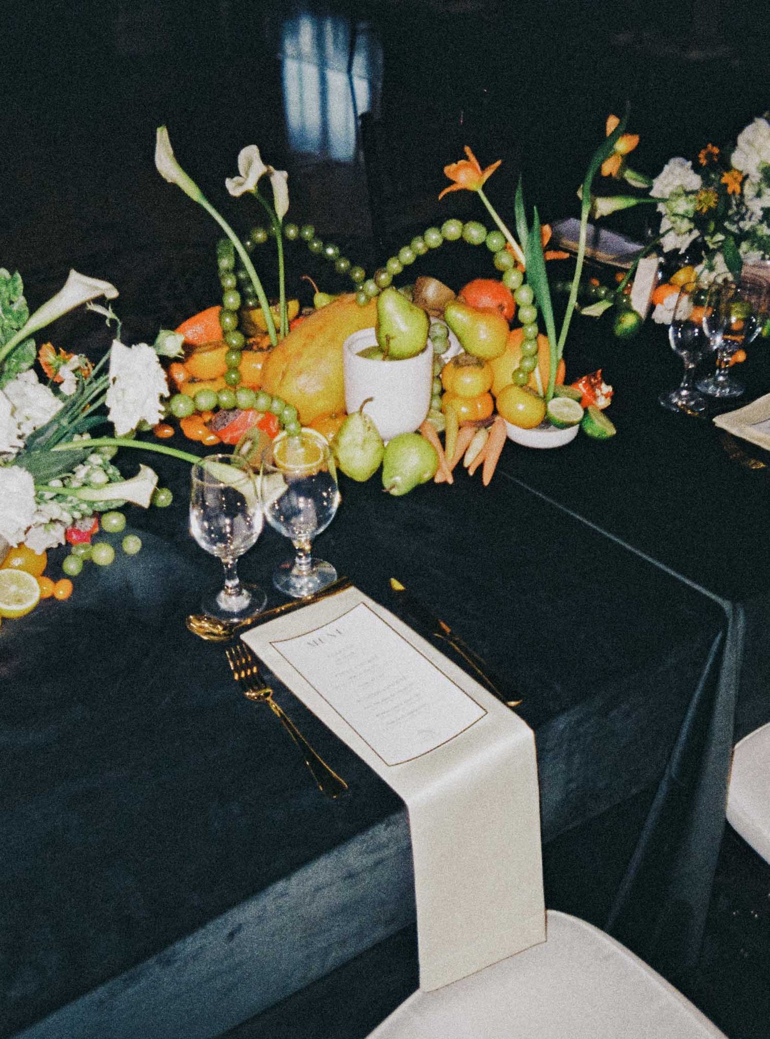 An elegant table setting with citrus fruits and floral centerpiece on a black tablecloth next to wine glasses and menu cards.