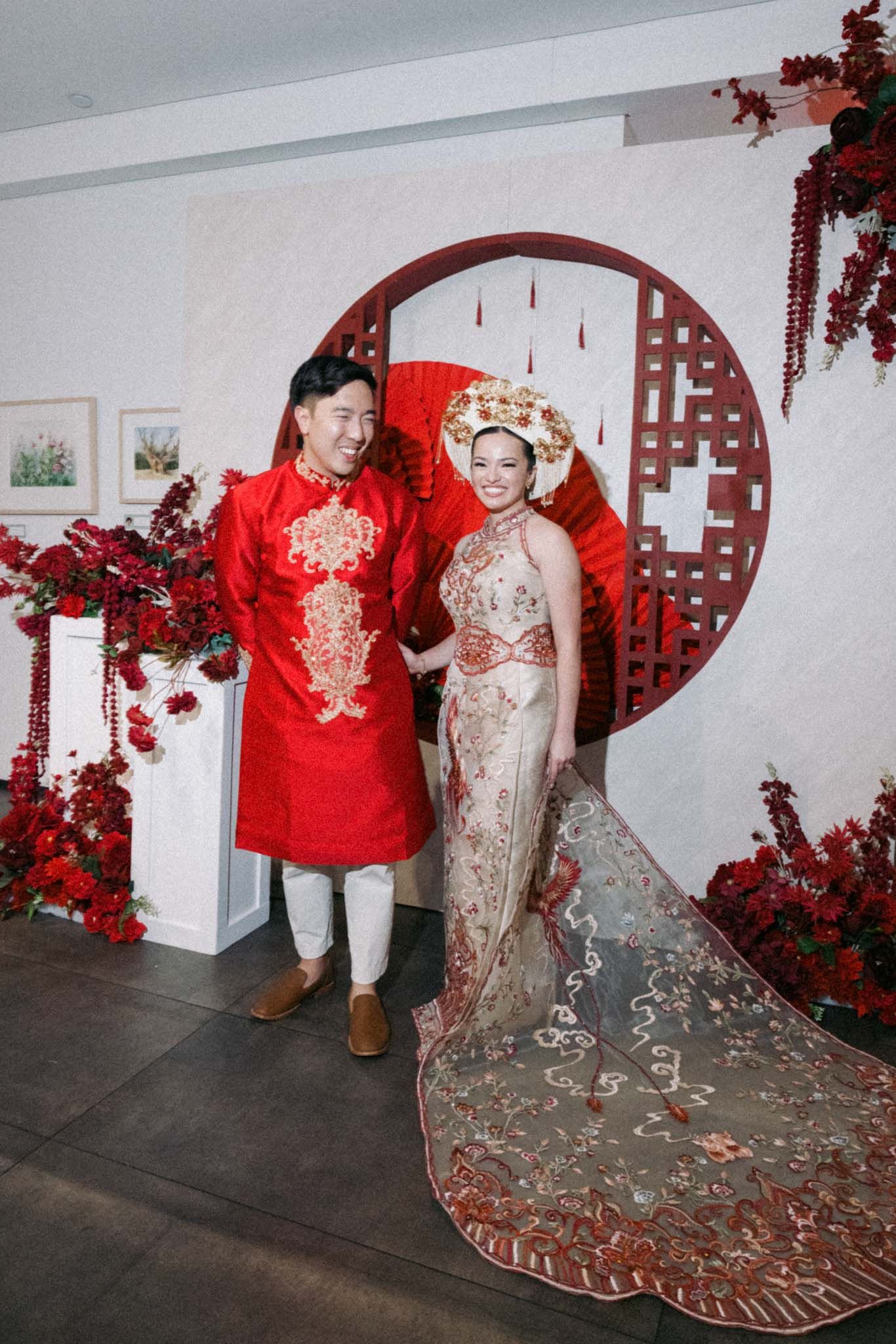 A couple in traditional Asian wedding attire poses in front of a red circular backdrop with cherry blossom decorations.