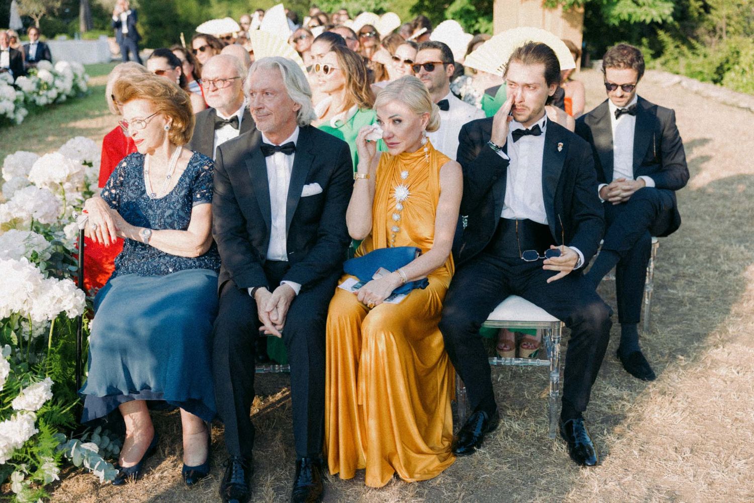 Wedding guests in formal attire gather outdoors for group photos during a sunny garden celebration.