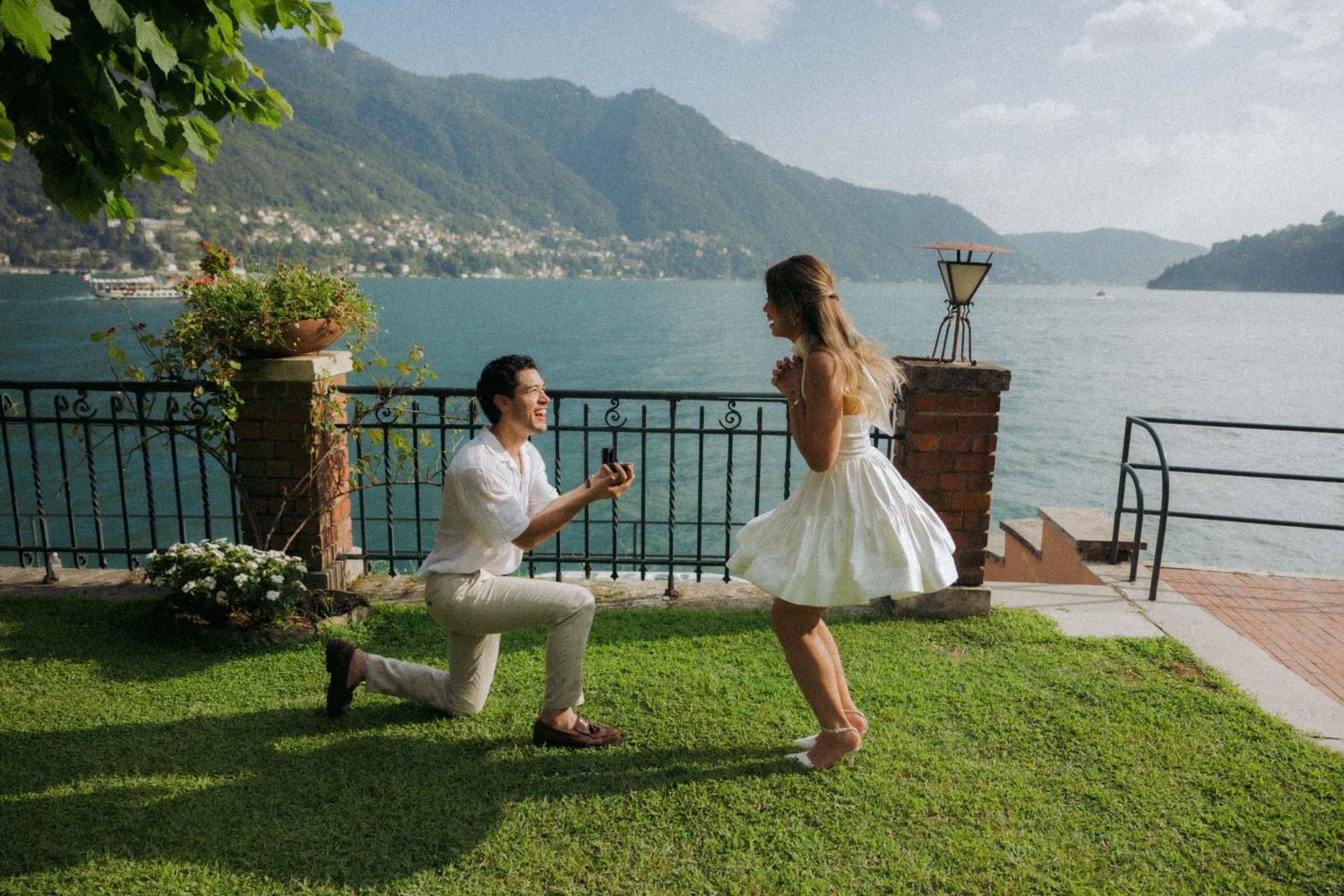Scenic view of a couple dancing on a grassy terrace overlooking a serene lake surrounded by mountains.
