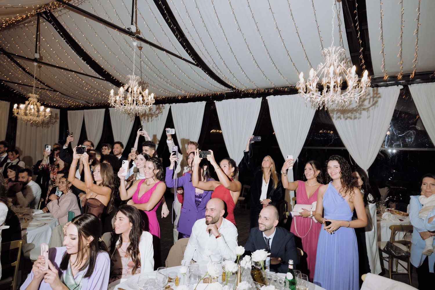 Wedding reception guests raise their hands in celebration under elegant crystal chandeliers in a draped tent venue.