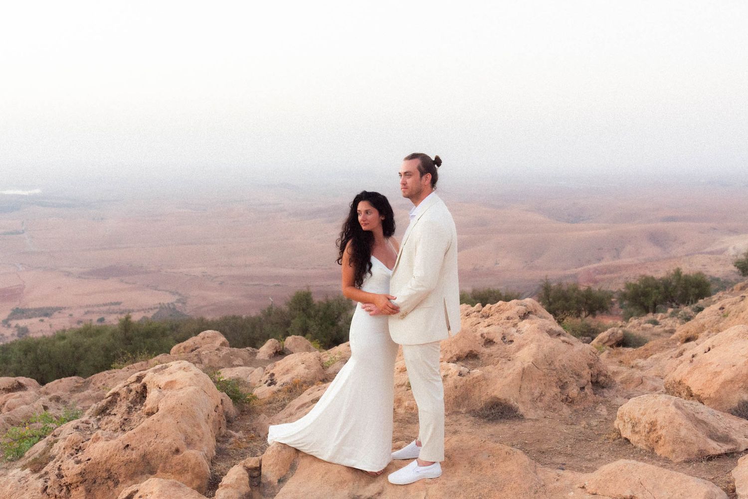 Two people in white outfits stand atop rocky desert cliffs overlooking a vast arid landscape at sunset.