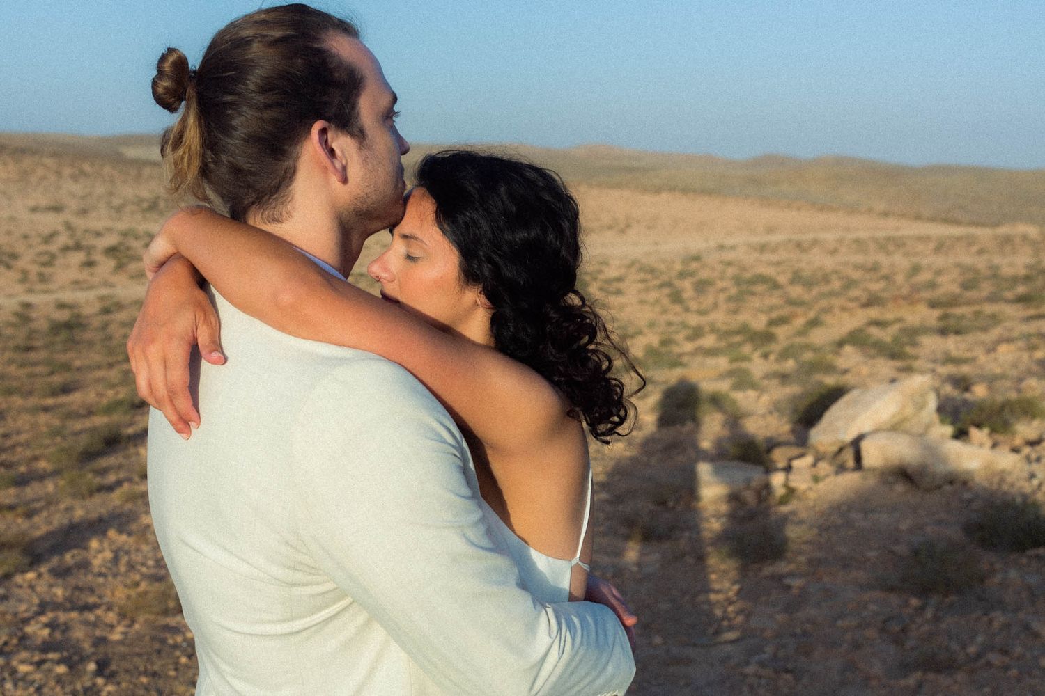 A couple embraces while standing in a desert landscape with mountains and open terrain in the background.