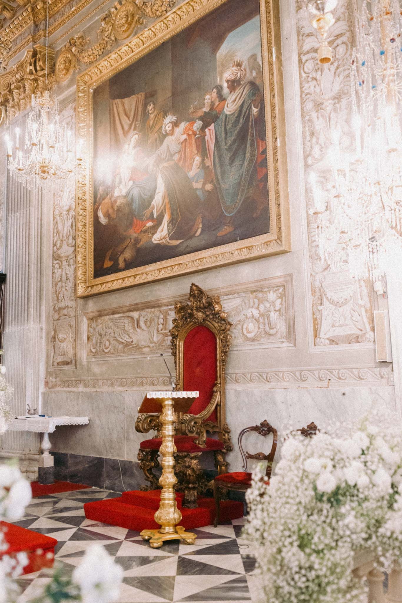 Ornate baroque church altar decorated with gold details, religious paintings, and white floral arrangements.