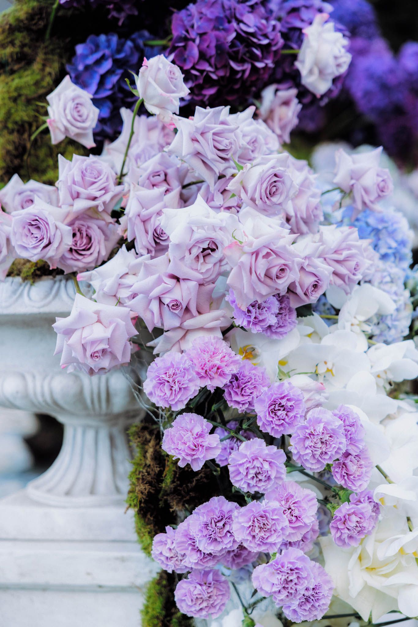 Beautiful floral arrangement featuring lavender roses and pink carnations cascading from a white pedestal with moss accents.