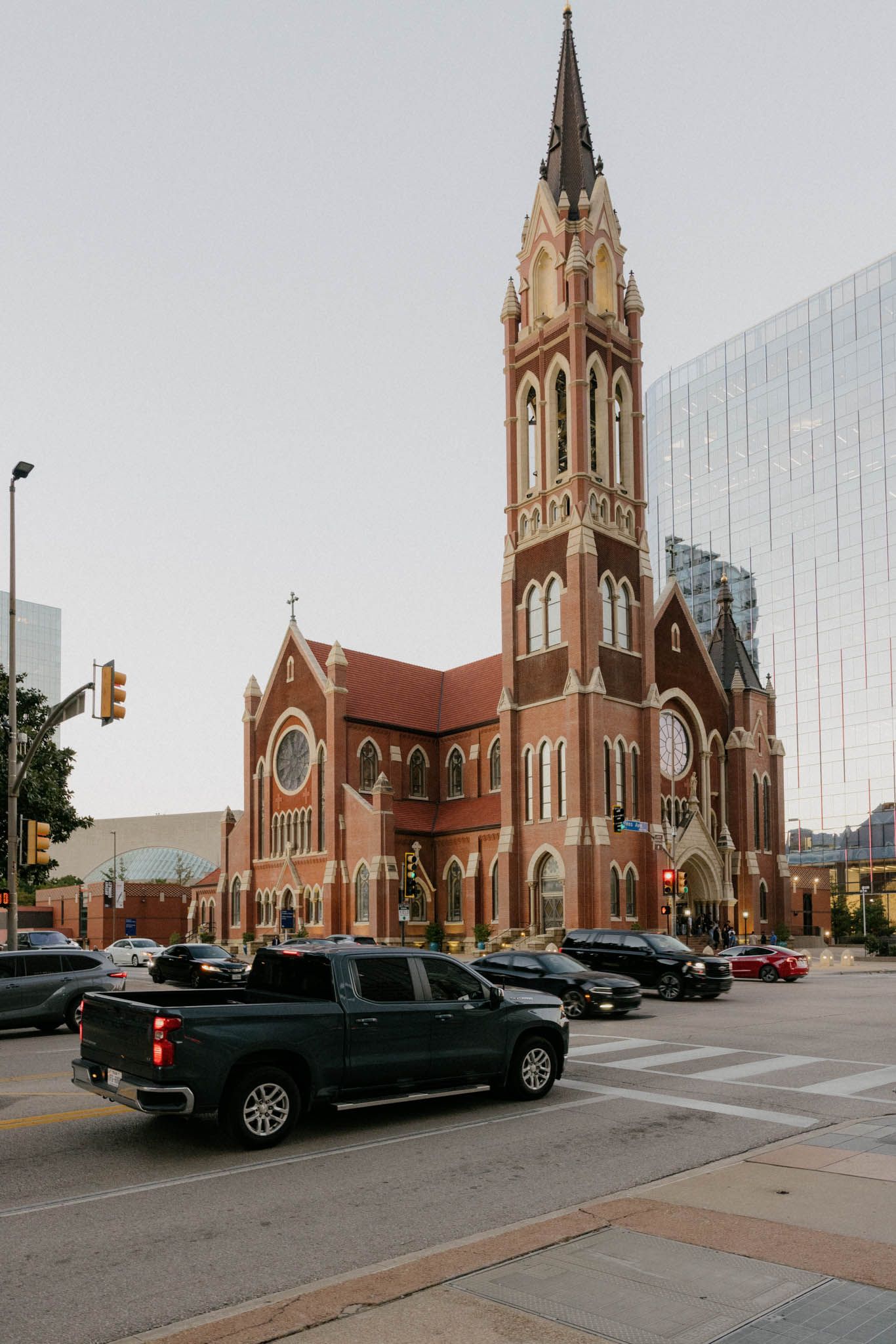Gothic revival church with red brick exterior and tall spires stands prominently on a city corner with parked cars nearby.