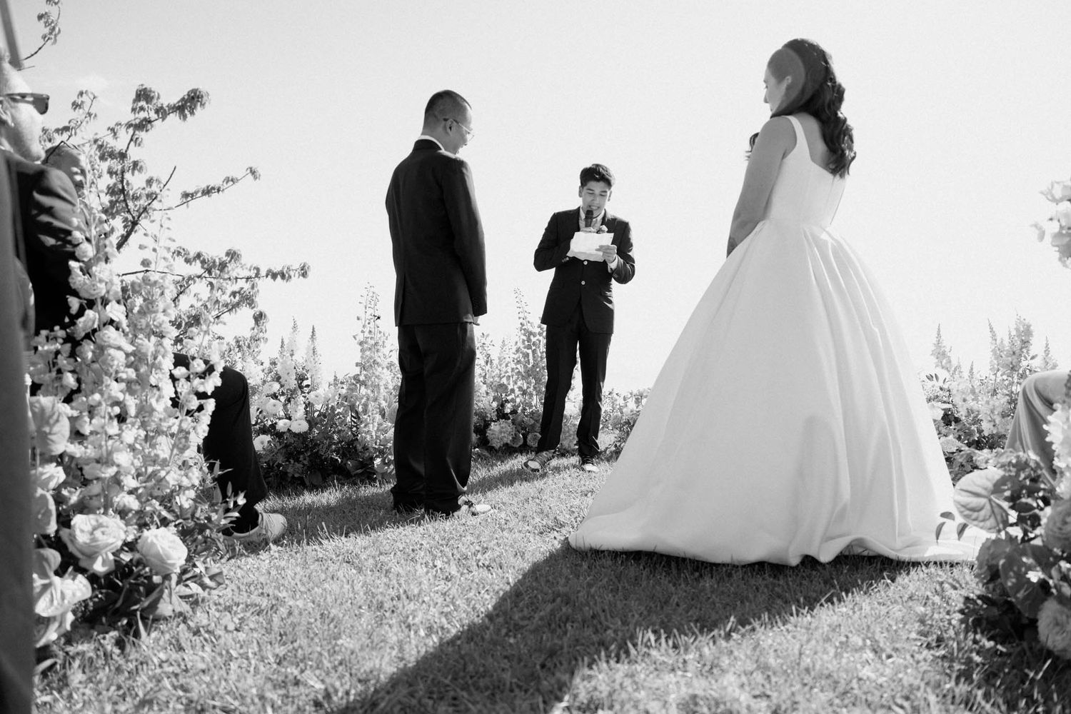 Wedding party stands on a grassy hillside in black and white silhouette against a bright sky.