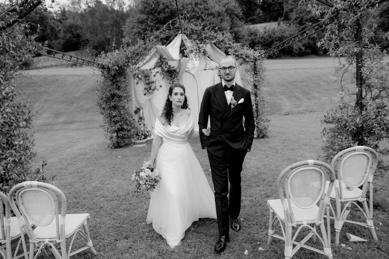 A bride and groom walk together down an outdoor garden aisle lined with elegant white chairs.