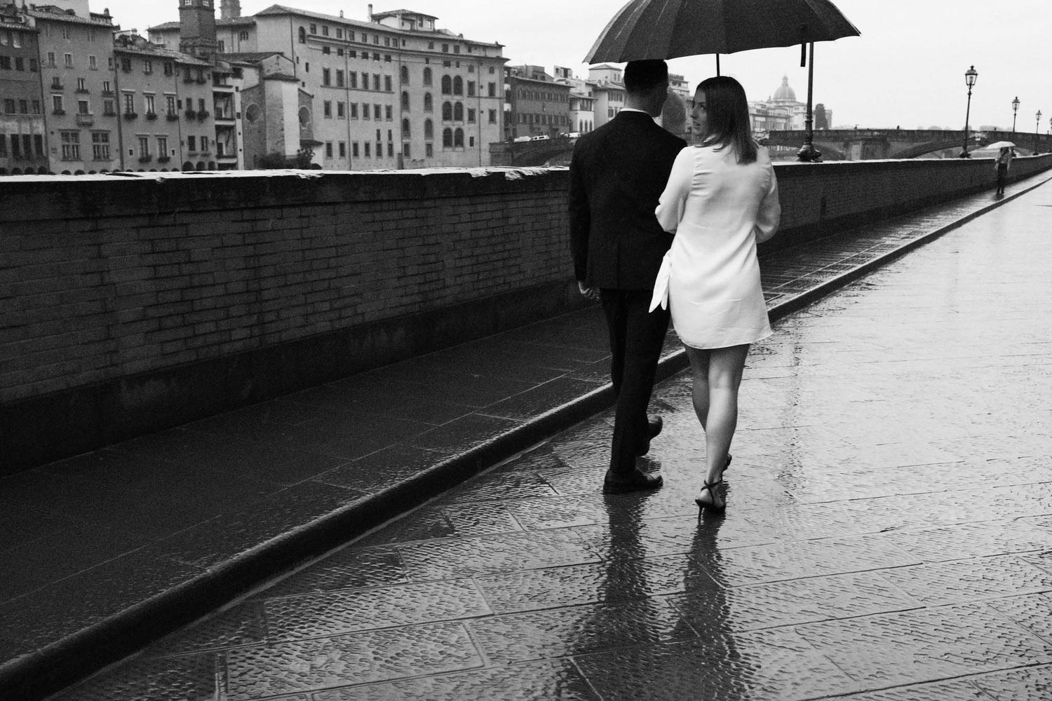 A couple walks together under a black umbrella on a rainy day along a brick wall in Florence, Italy.