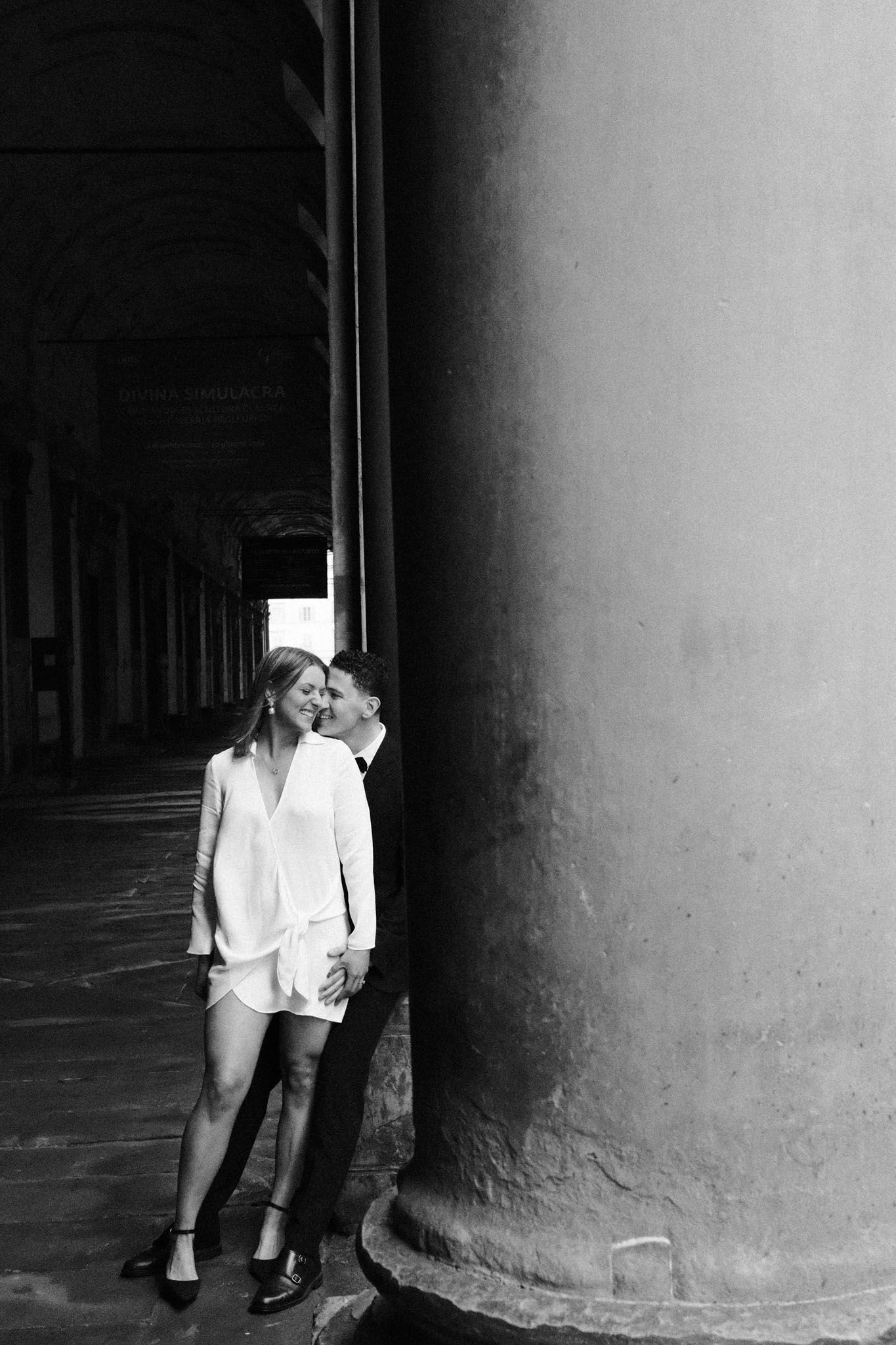 A couple shares an intimate moment behind a large stone column in a dimly lit corridor.