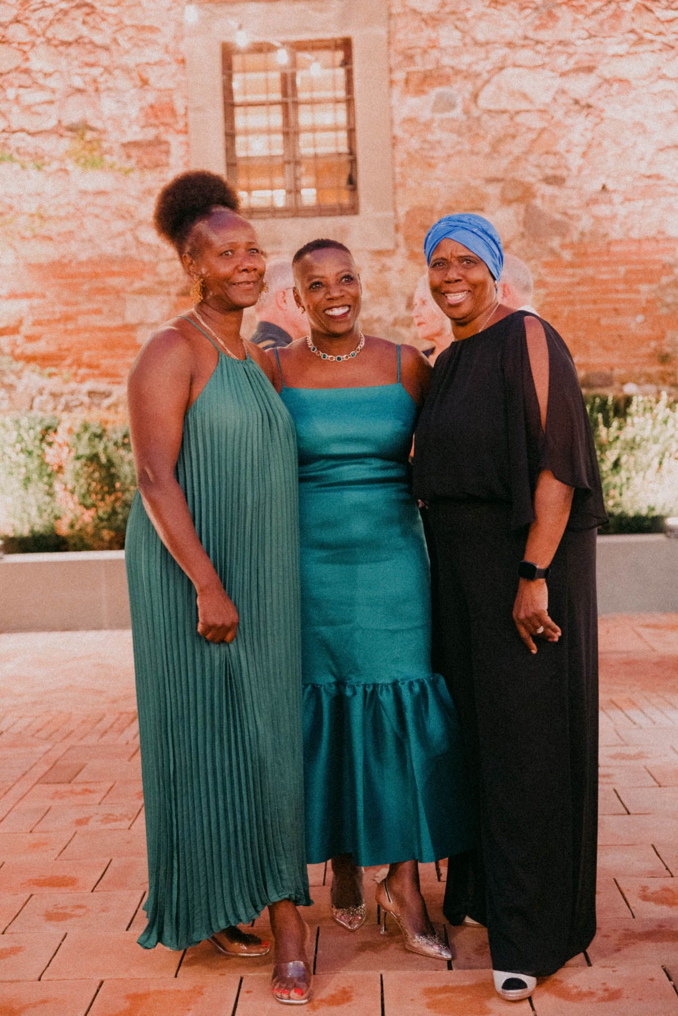 Three women pose together in elegant evening wear including green pleated and teal fitted dresses against a rustic brick wall.
