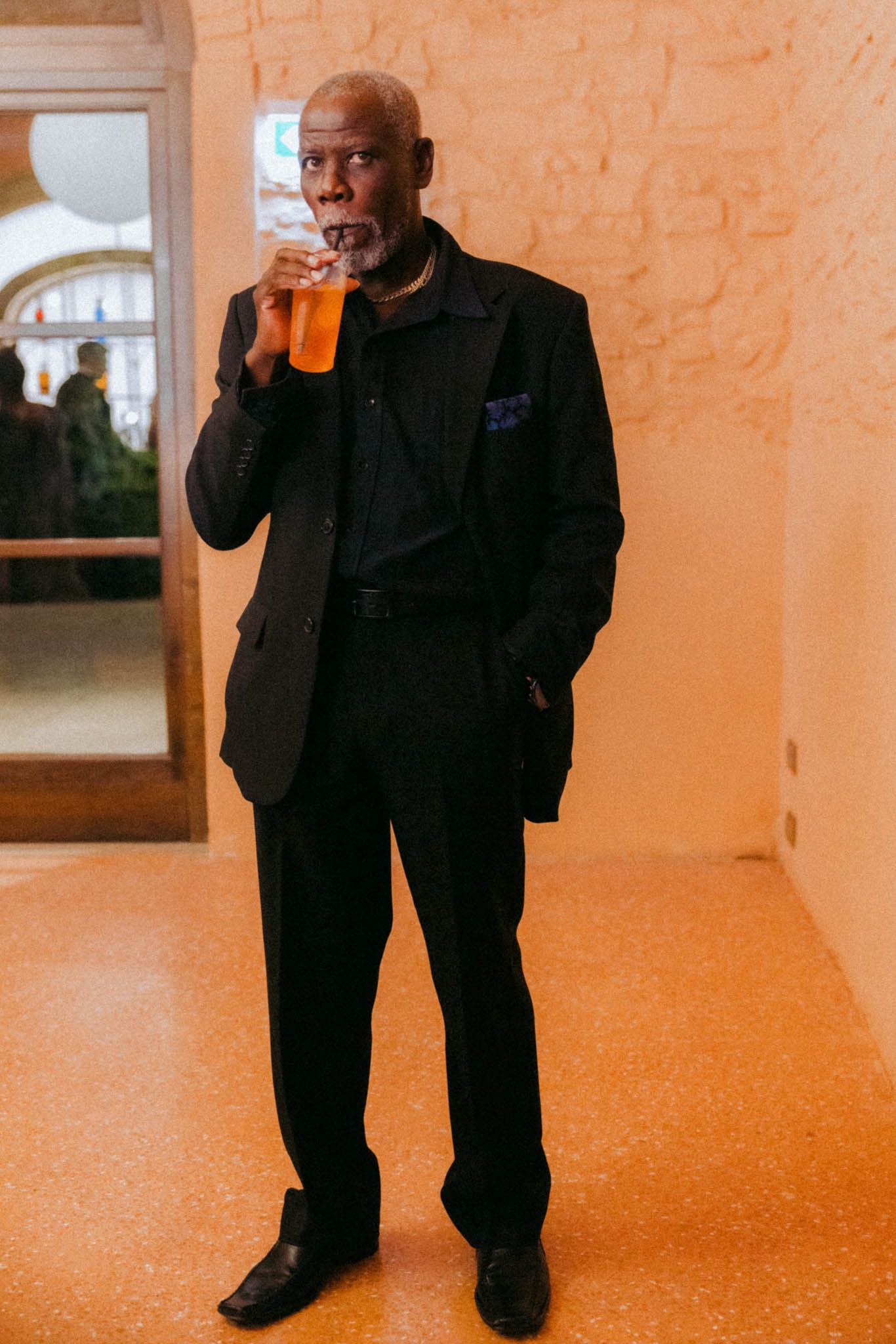 A person wearing all black sits and drinks from a glass in an orange-tinted room.