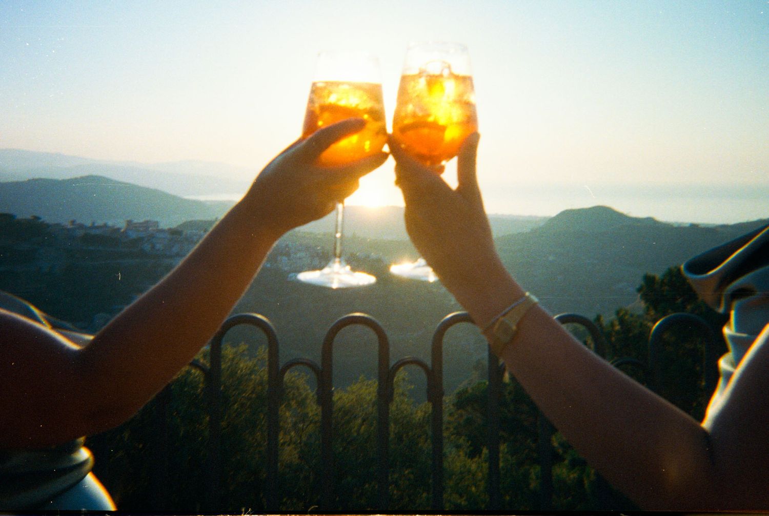 Hands holding up a glass of wine against a sunset mountain landscape, creating a golden silhouette effect.