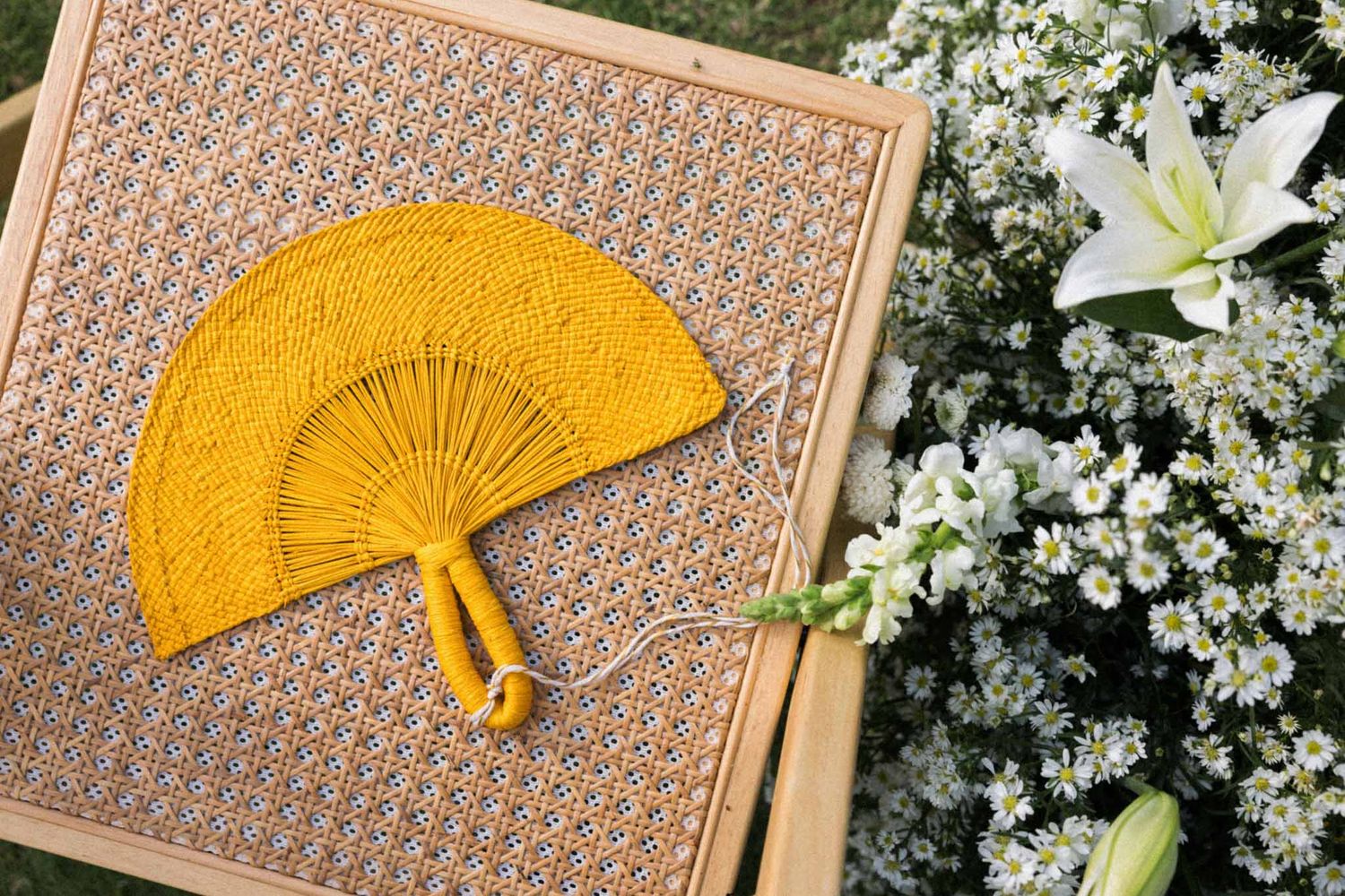 A bright yellow lace hand fan rests on a wooden surface next to white lily and small white flowers.